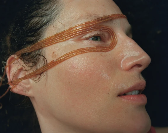 Close-up of woman's face with orange beaded strands wrapped around her head, side profile against a dark background.
