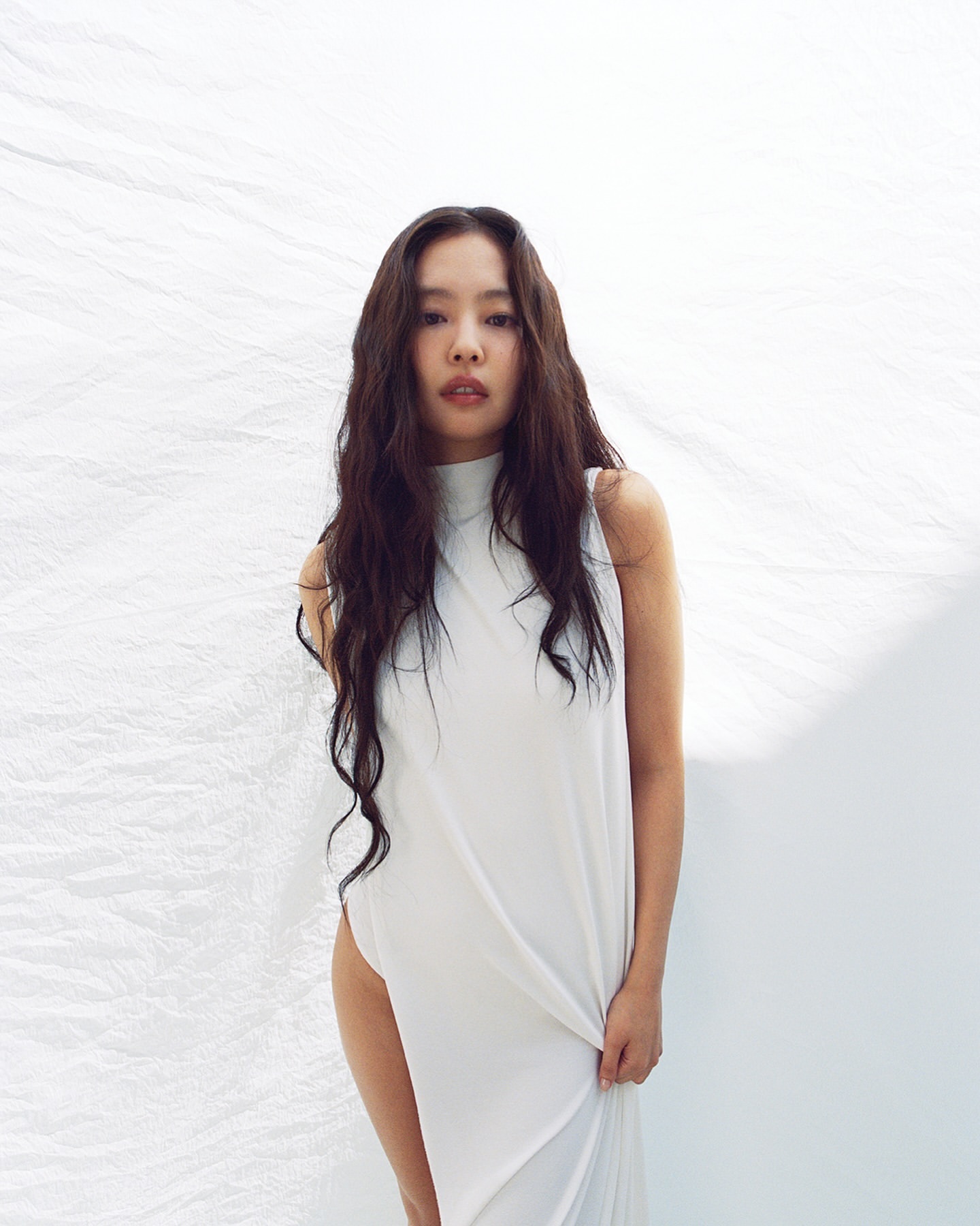 Portrait of a woman with long dark hair wearing a white dress standing in front of a white fabric backdrop in a photo studio.