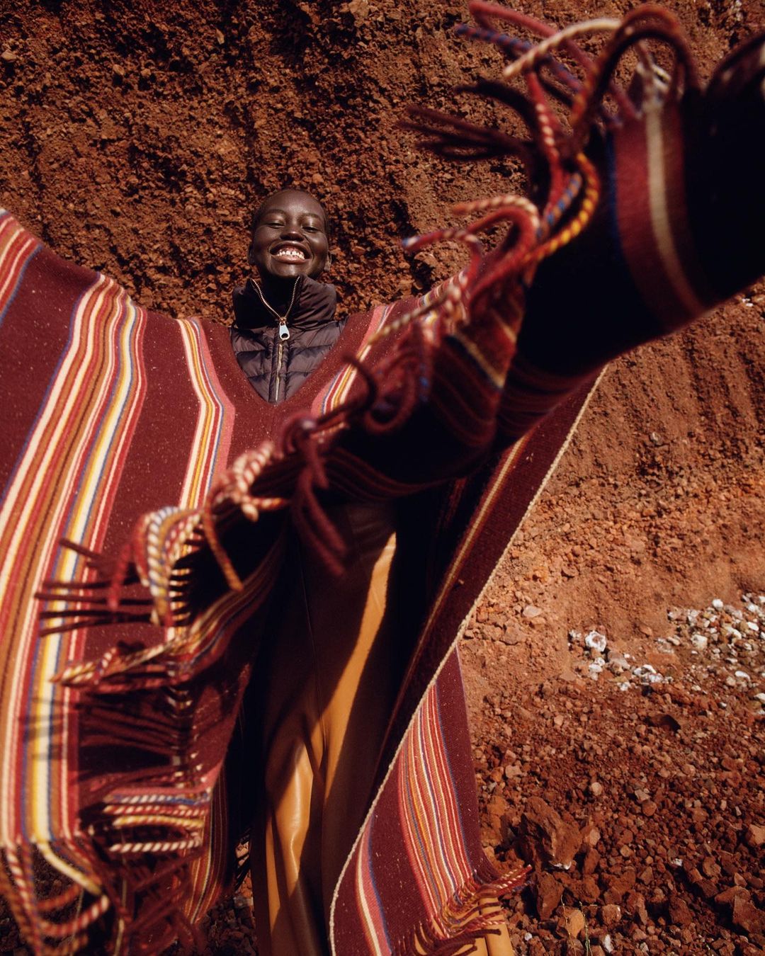 Smiling African woman in poncho standing against red clay backdrop, outdoor fashion shot with vibrant colors and textures.