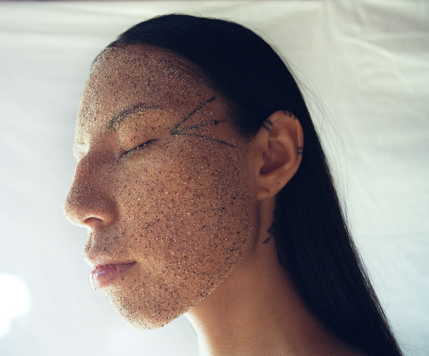 Profile of woman's face covered in sand, eyes closed, neutral expression in studio with a neutral, soft light and white background.