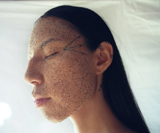Profile of woman's face covered in sand, eyes closed, neutral expression in studio with a neutral, soft light and white background.