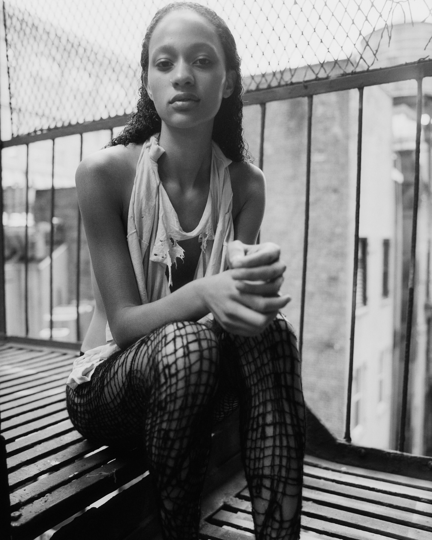 Black and white portrait of an adult female with curly hair sitting on metal stairs in fishnet leggings with buildings in the background.