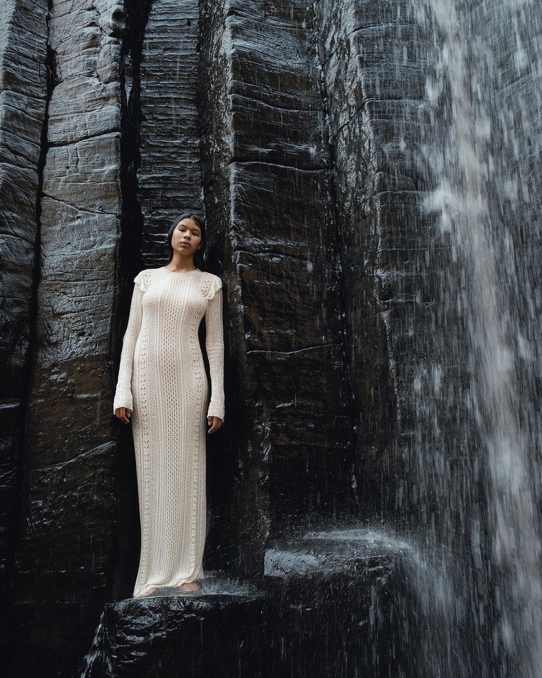 Woman in long cream dress poses on rocks near dark rockface with waterfall. Full-length shot of model in a natural setting.