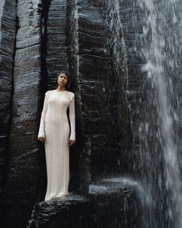 Woman in long cream dress poses on rocks near dark rockface with waterfall. Full-length shot of model in a natural setting.