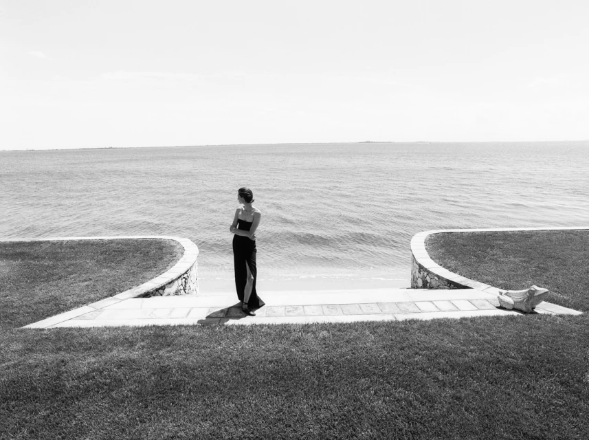 Woman in black dress stands on a stone pathway near the ocean on a sunny day. Lush grass and a seaside view create a serene setting.