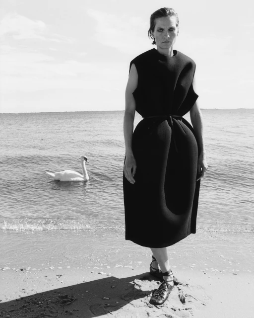 Black and white fashion portrait of a woman in a black dress standing on a beach next to the water with a swan swimming in the sea.