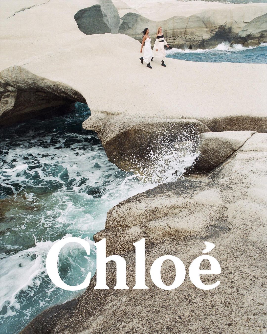Two women in white dresses walk on rocky coastline by the ocean with waves. Fashion advertising campaign by Chloé.