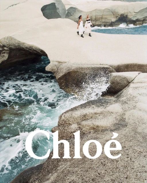 Two women in white dresses walk on rocky coastline by the ocean with waves. Fashion advertising campaign by Chloé.
