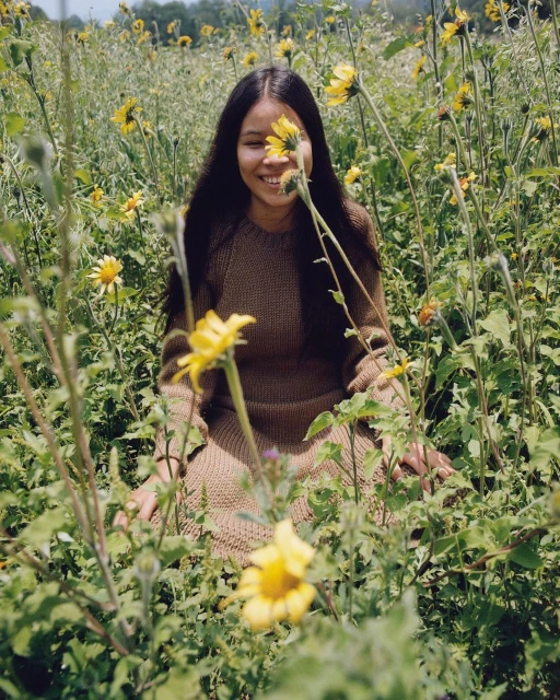 Smiling woman in a field of yellow wildflowers on a sunny day, wearing a brown sweater, enjoying nature in an outdoor setting.