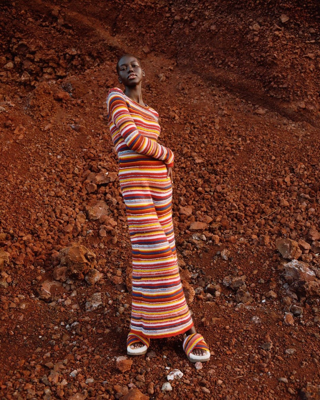 A full-shot fashion portrait of an adult woman in a colorful striped two-piece outfit, posing outdoors against a red rocky background.