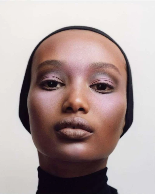 Close-up studio portrait of an adult female with dark skin wearing a black head covering, showcasing mauve makeup and a neutral facial expression.