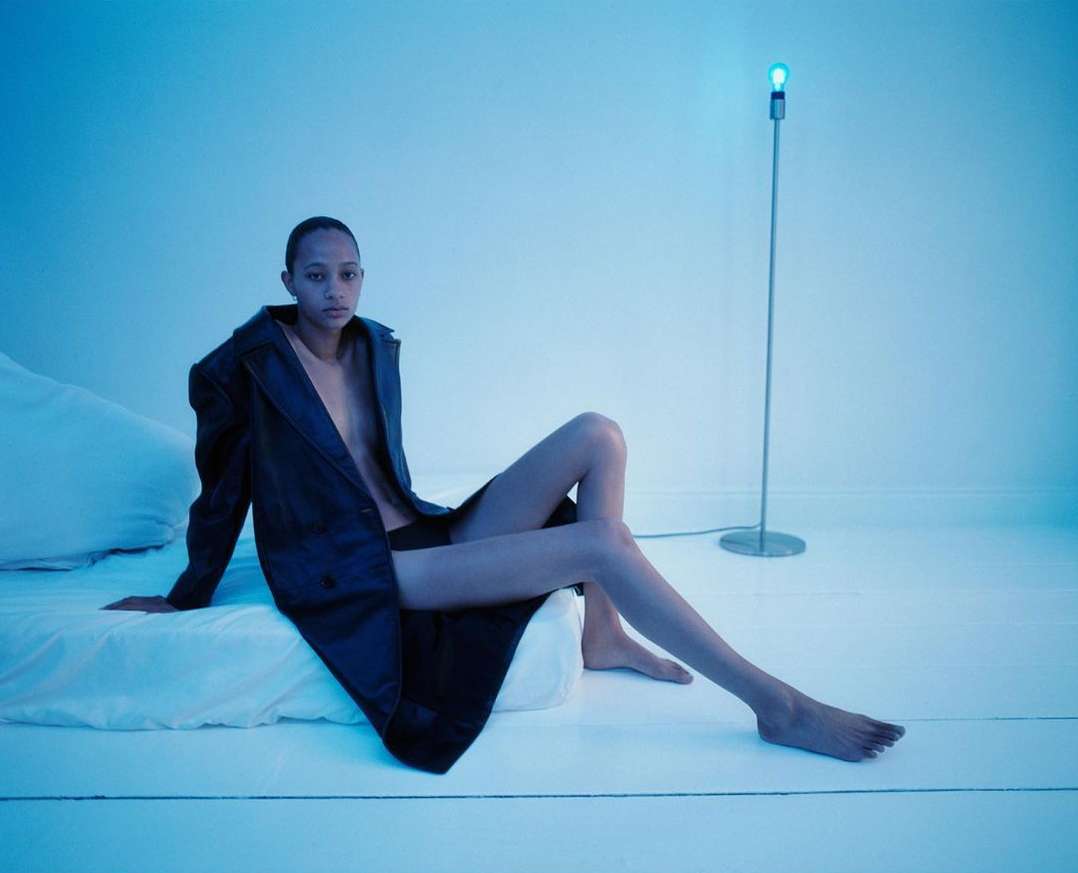 A person poses on a white bed with a black coat, illuminated by a modern floor lamp in a minimalist studio setting with blue lighting.