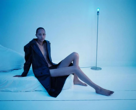 A person poses on a white bed with a black coat, illuminated by a modern floor lamp in a minimalist studio setting with blue lighting.