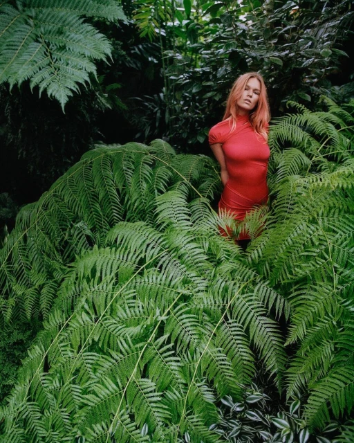 A young woman with red hair stands amidst lush green ferns in a natural setting, wearing a red dress, in a verdant, immersive, natural environment.