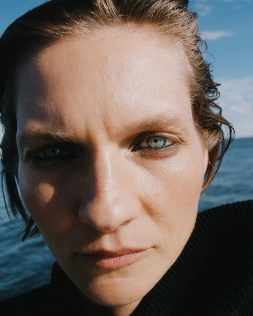 Close-up portrait of an adult woman with wet, short dark hair and blue eyes against an outdoor backdrop of the ocean and blue sky.