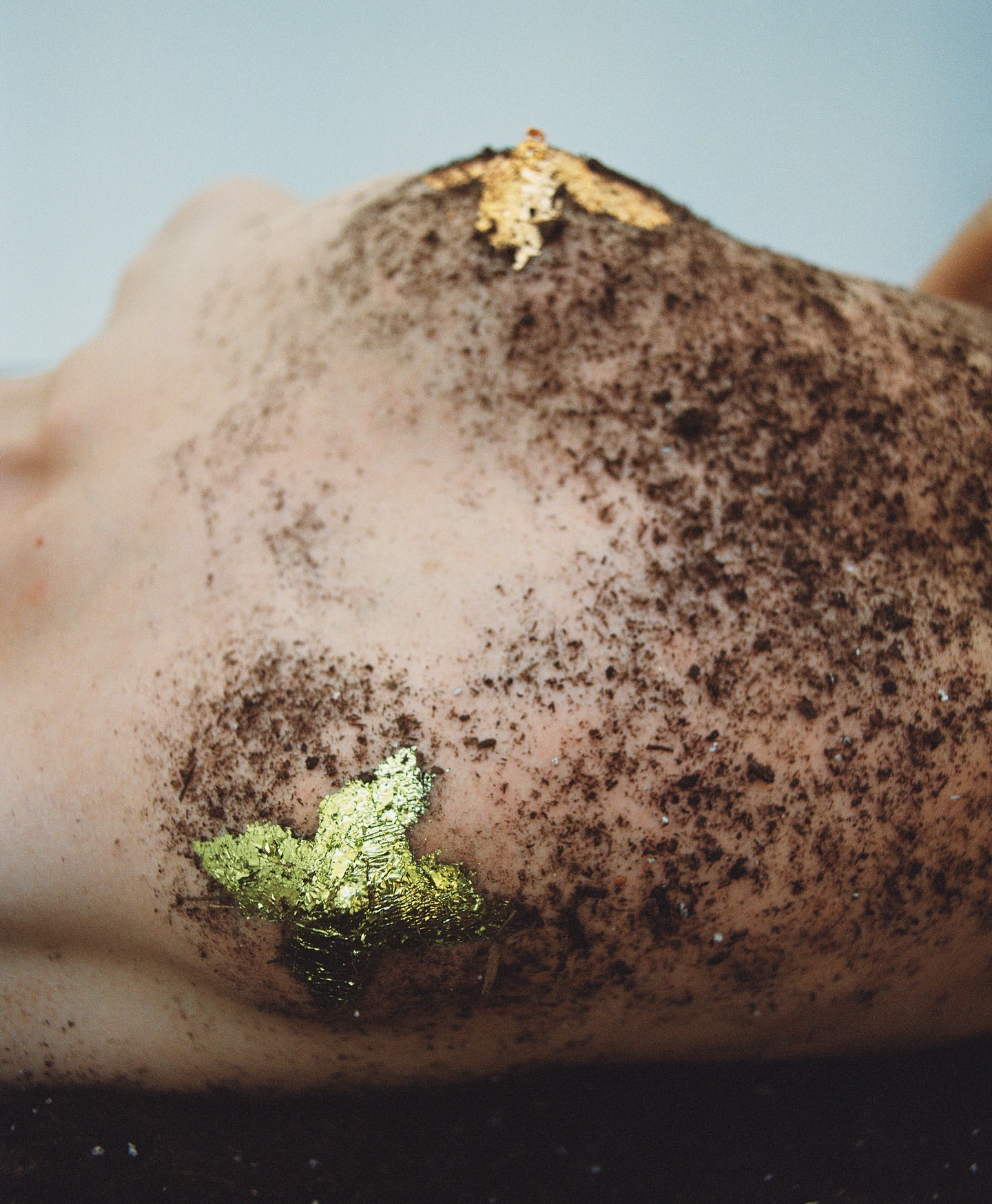 Close-up of an adult lying on a dark surface, with soil and gold flakes covering part of their body against a light background.