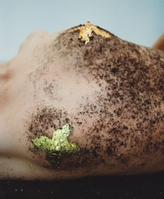 Close-up of an adult lying on a dark surface, with soil and gold flakes covering part of their body against a light background.