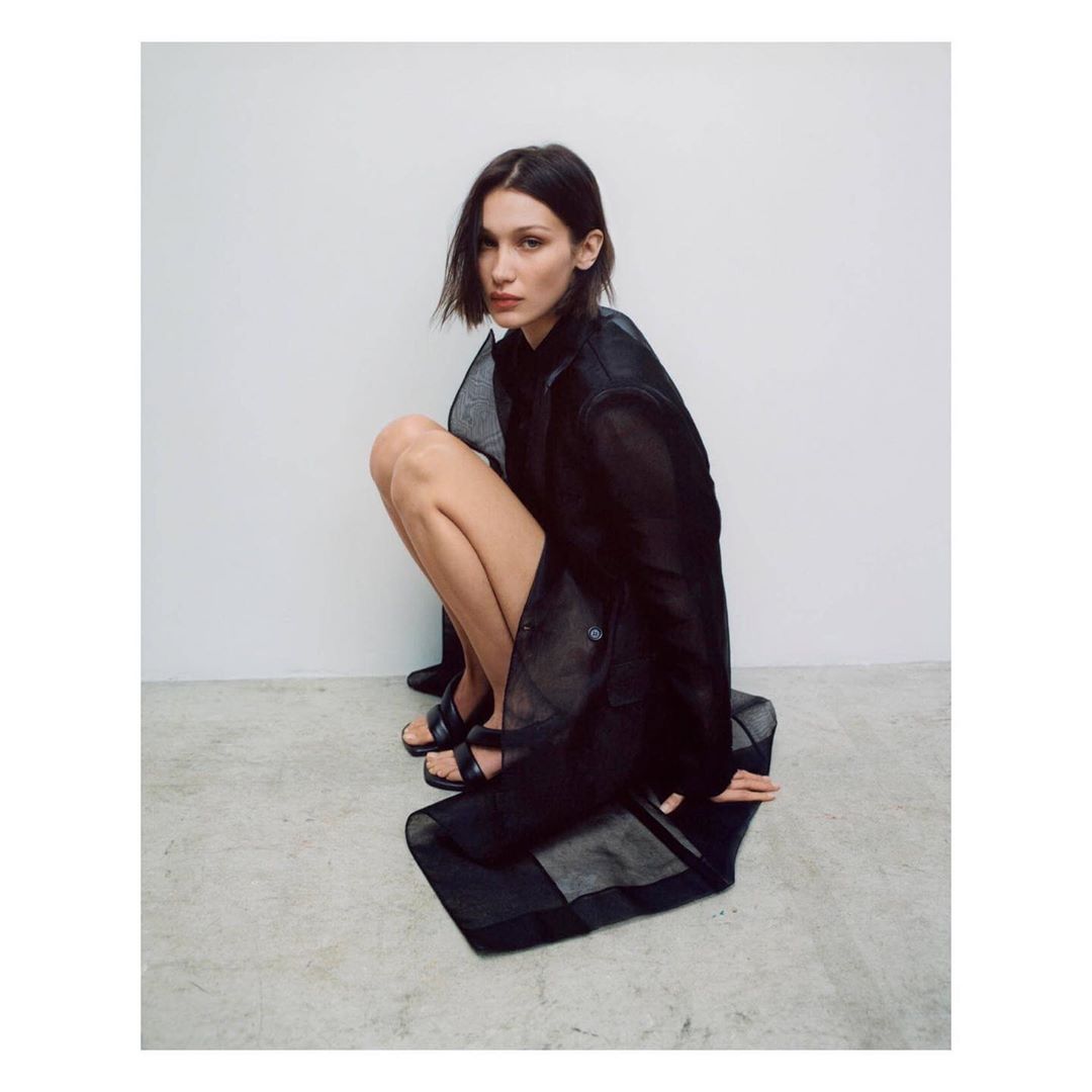 A young adult woman with brown hair is crouched indoors wearing a sheer black jacket and black shoes in a fashion editorial shot.