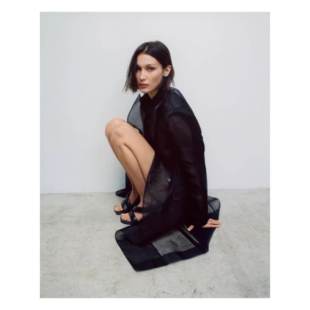 A young adult woman with brown hair is crouched indoors wearing a sheer black jacket and black shoes in a fashion editorial shot.