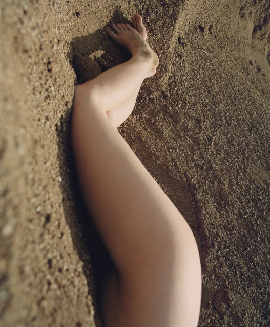 Woman's legs partially buried in sand at the beach, illuminated by soft sunlight. The focus is on the texture of the sand.