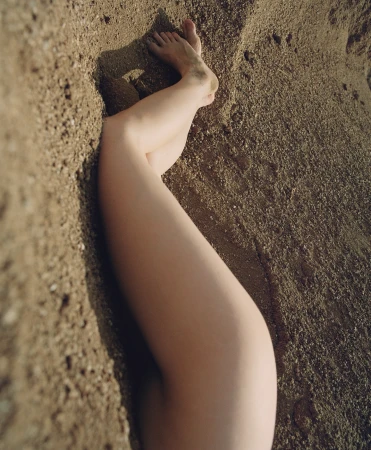 Woman's legs partially buried in sand at the beach, illuminated by soft sunlight. The focus is on the texture of the sand.