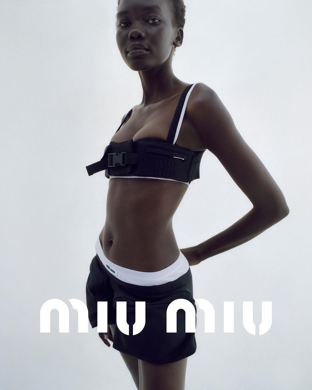 Dark-skinned woman poses in a black bralette and black skirt with white trim against a white background, fashion studio shot.