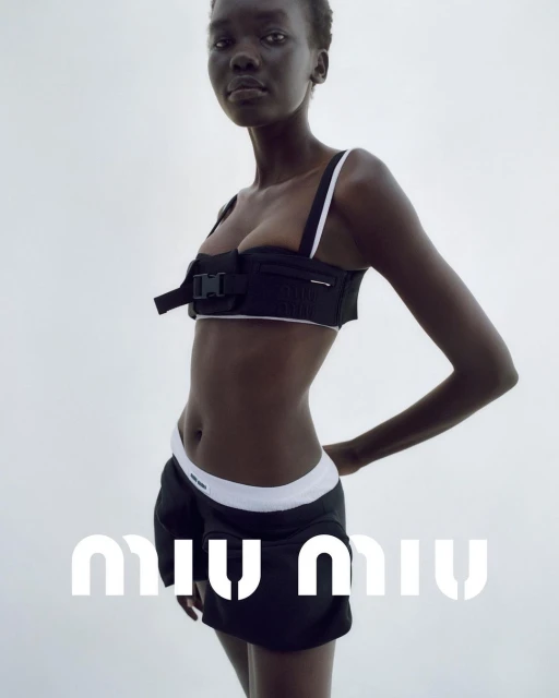 Dark-skinned woman poses in a black bralette and black skirt with white trim against a white background, fashion studio shot.