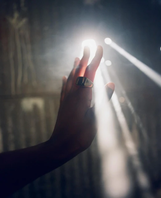 An adult hand adorned with a gold ring reaches up to the bright light, with beams of light shining in the background, indoor setting.
