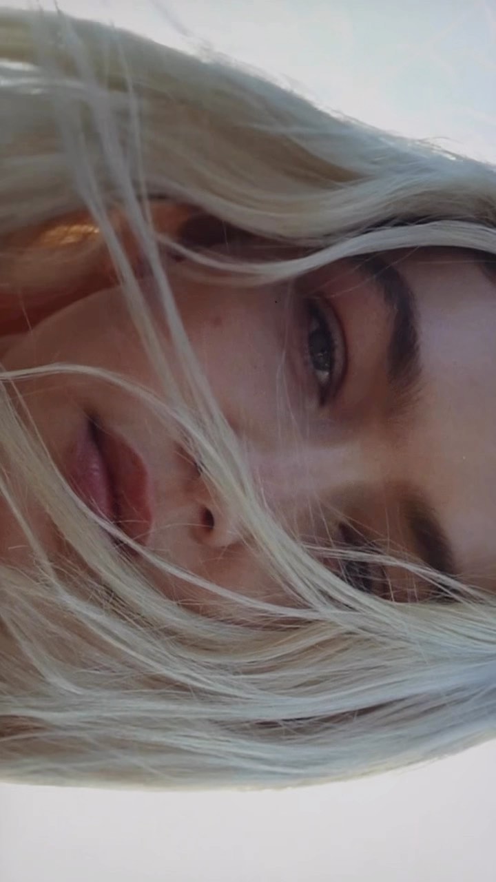 Close-up portrait of a blonde woman with loose hair blowing across her face against a bright sky background.