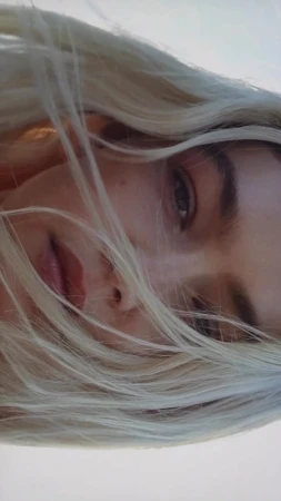 Close-up portrait of a blonde woman with loose hair blowing across her face against a bright sky background.