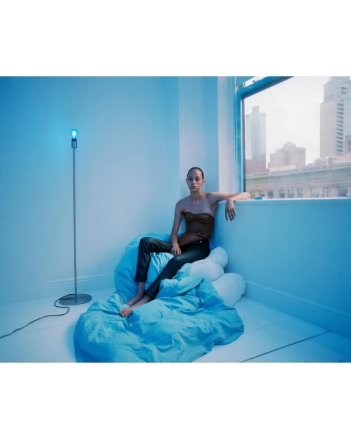 A woman poses near a window in a studio apartment with a blue color scheme, wearing a brown top and dark pants, illuminated by a floor lamp.