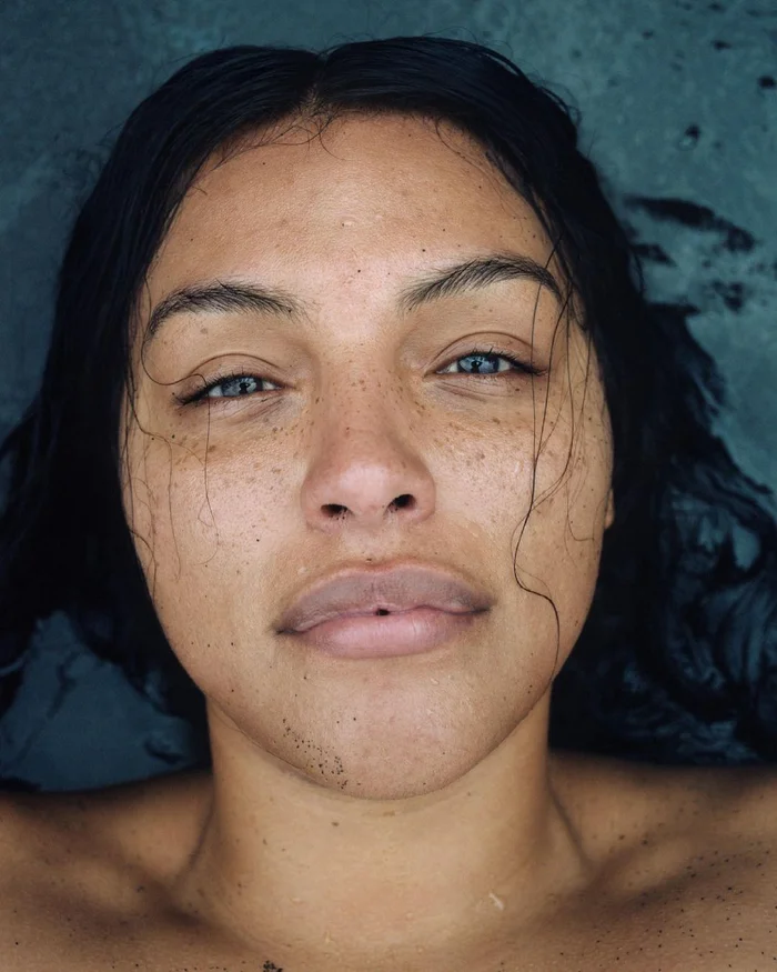 Close up portrait of a woman with wet dark hair and freckles against a dark stone background photographed by Zoe Ghertner
