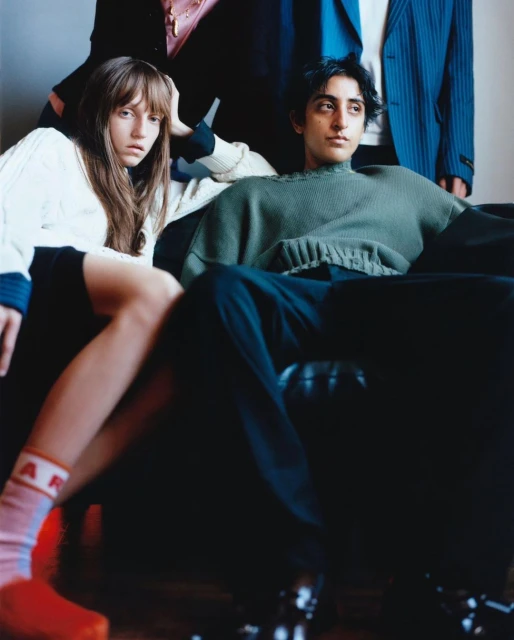Group of four people in indoor setting. Three are partially visible, while one man sits on a black chair. One woman stands.