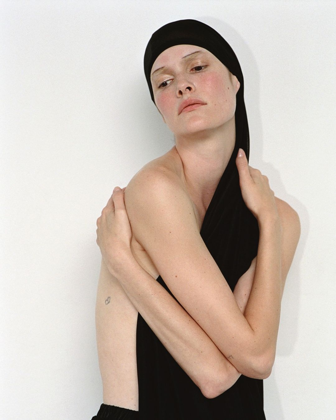 Portrait of a fair-skinned person with a black head covering, arms crossed, looking down against a neutral wall in a bright studio setting.