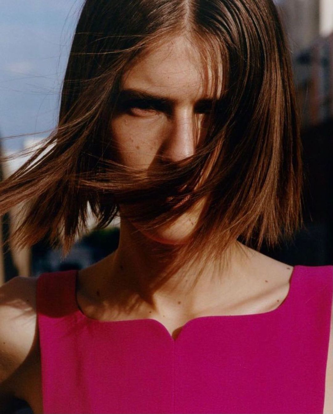 Close-up portrait of a young adult woman with short brunette hair, wearing a pink dress, in an urban outdoor setting with diffused natural light.