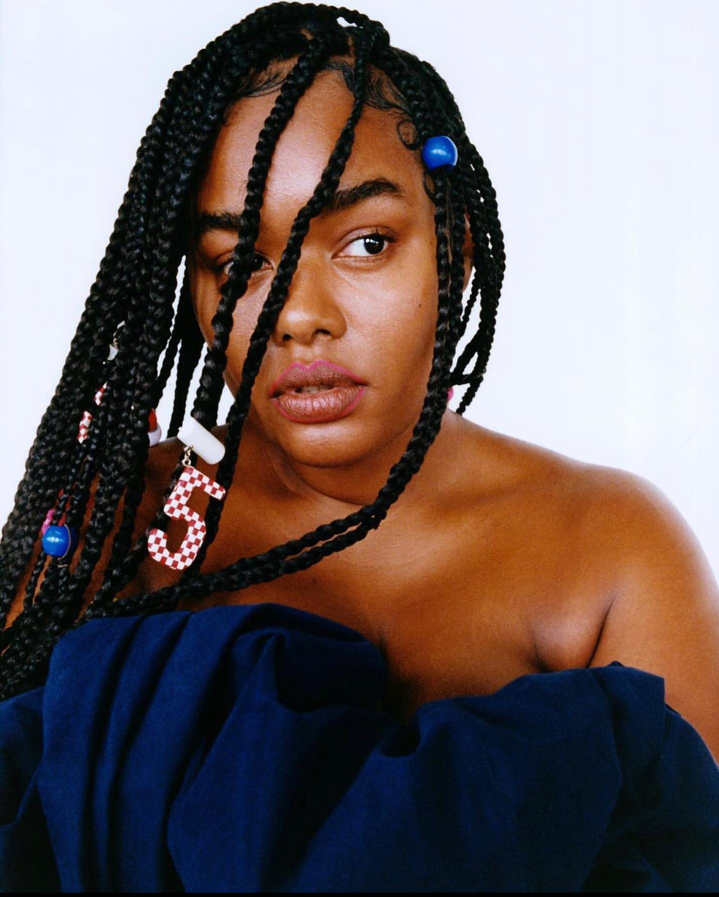 Portrait of a Black woman with braided hair, blue ruffled top, and playful accessories, posing against a clean white backdrop in a studio.