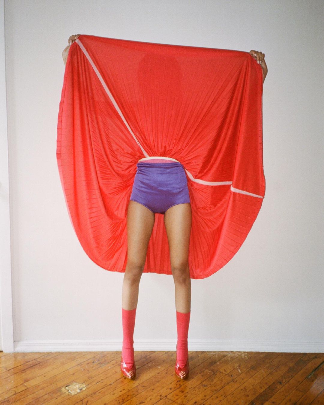 Person holding up a large pleated red fabric covering their upper body, wearing purple high-waisted shorts and red socks.