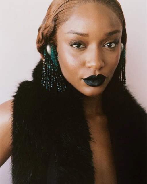 Close-up portrait of a stylish African-American woman with black lipstick, a fur vest, and beaded earrings in a neutral setting.