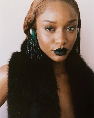 Close-up portrait of a stylish African-American woman with black lipstick, a fur vest, and beaded earrings in a neutral setting.