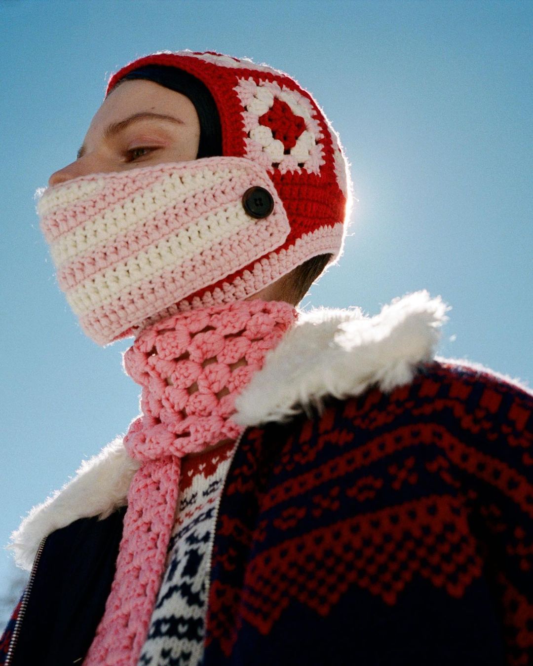 Person wears a red and white crochet balaclava with a pink scarf and fair isle sweater against a clear blue sky in a portrait shot.