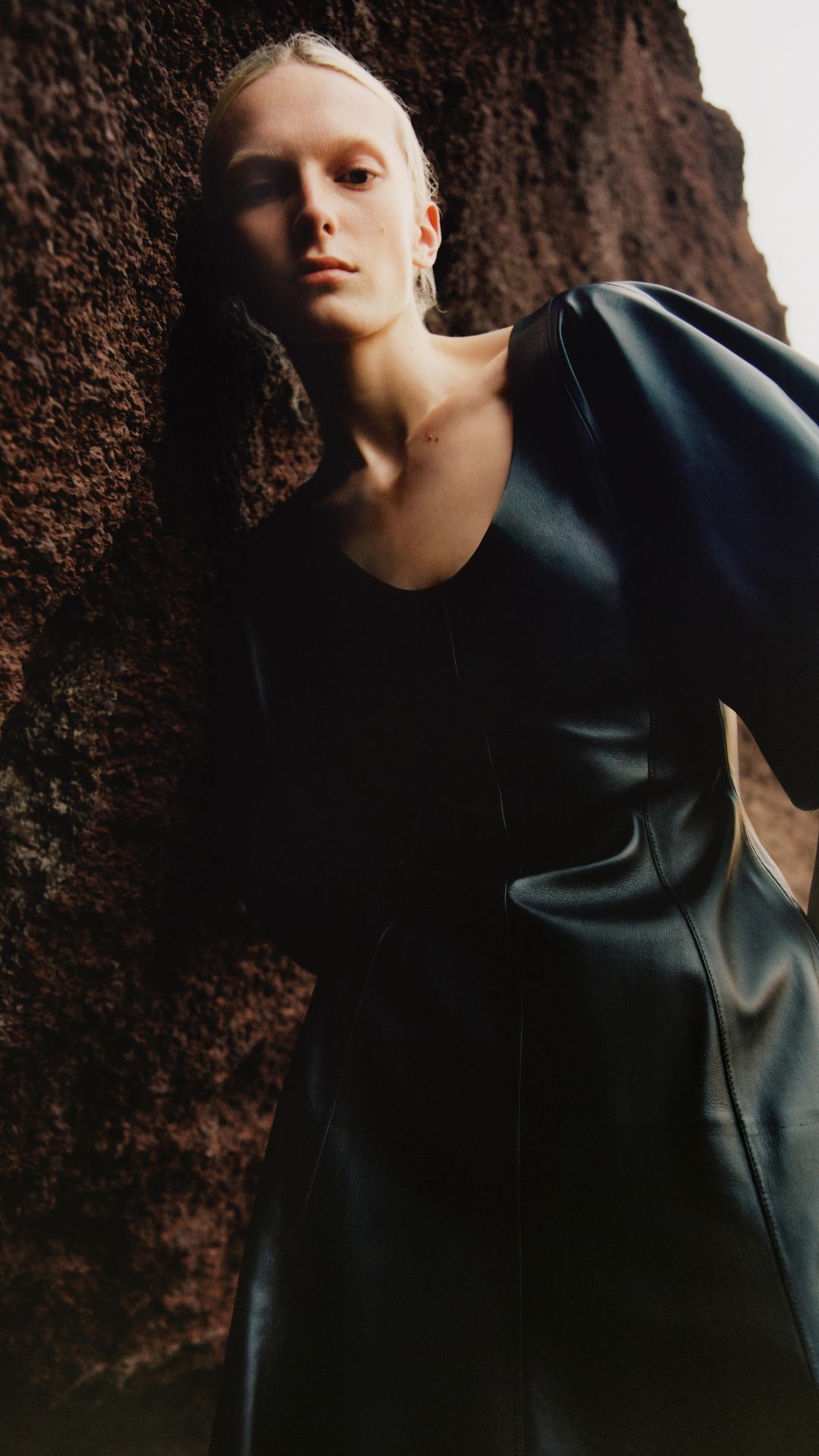A fair-skinned woman with blonde hair poses in a black dress, next to a textured brown surface, with diffused lighting.