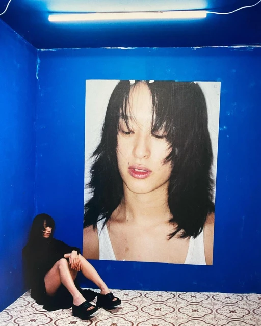 Portrait of a young Asian woman sitting in a blue corner room next to a poster of herself in black and white, fashionable and cool.