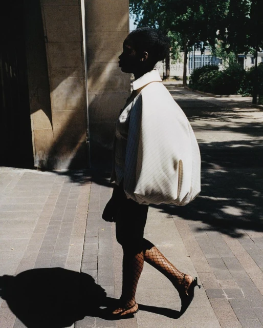 A silhouetted woman with dark skin walks on a city sidewalk in heels and fishnets wearing a stylish white jacket with puffy shoulders.