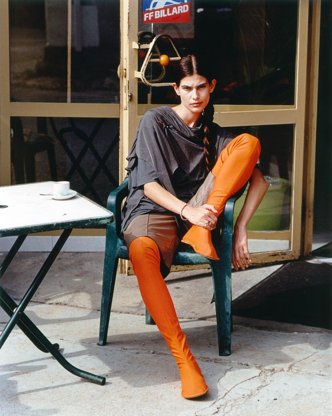 A fashionable adult female with braided hair, wearing bright orange thigh-high boots, poses in a gray top and brown shorts on an outdoor patio.