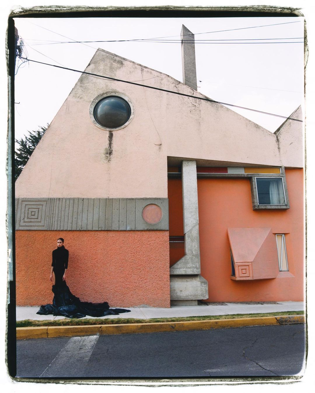 Woman in black outfit stands next to modern architectural building with geometric elements, soft colors, and circular window detail.