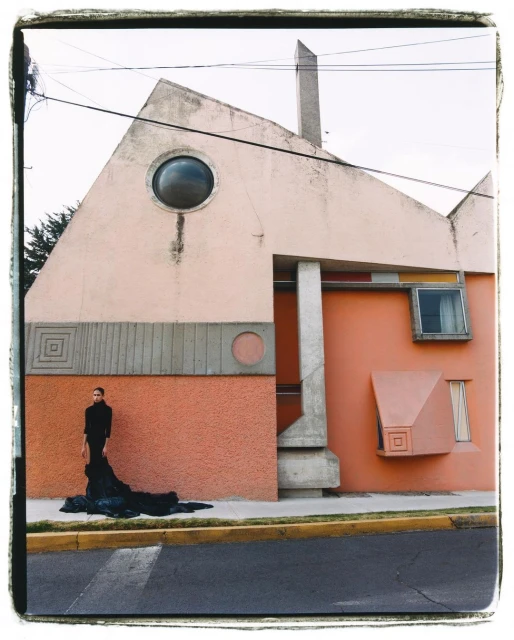 Woman in black outfit stands next to modern architectural building with geometric elements, soft colors, and circular window detail.