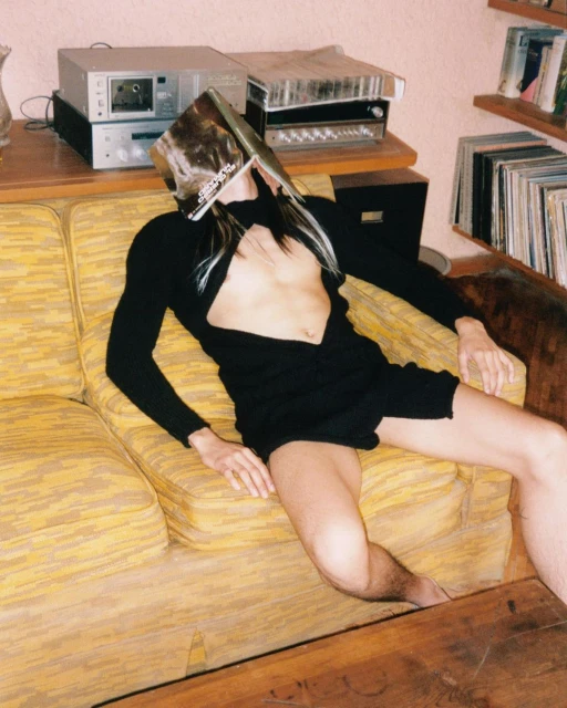 A person in a black cutout dress reclines on a couch with a magazine over their face, surrounded by vintage electronics and vinyl records.