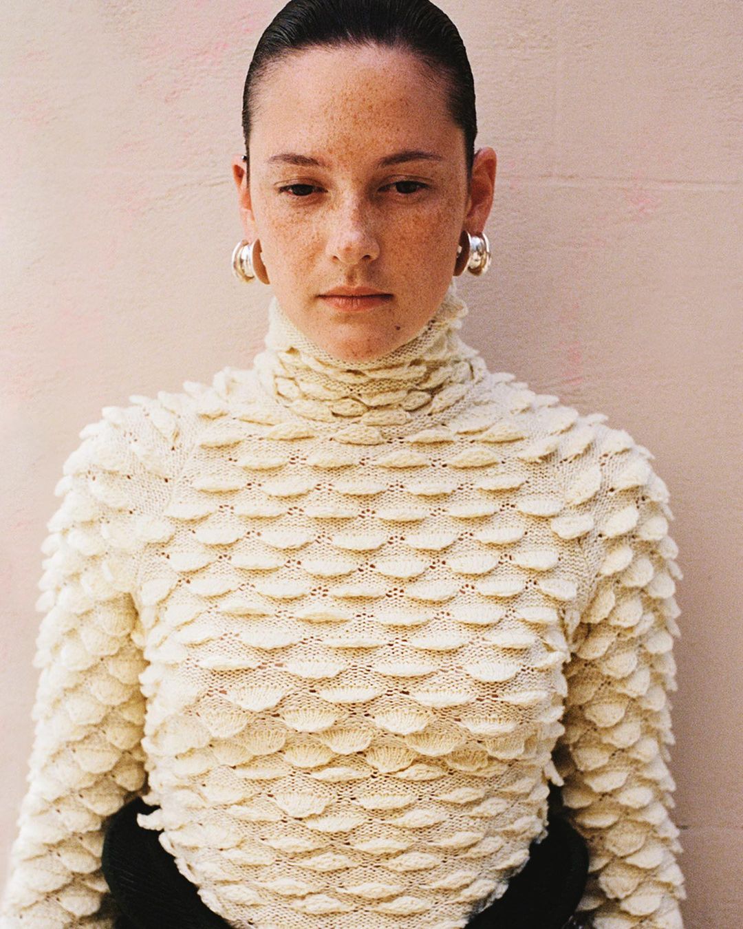 Close-up shot of a freckled woman with slicked-back hair wearing a textural ivory turtleneck and silver hoop earrings against a pink wall.
