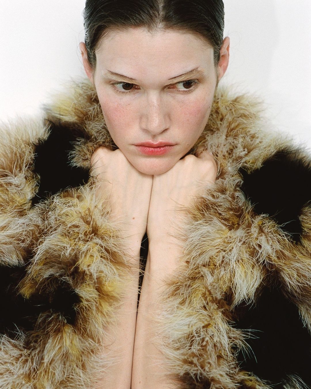 Portrait of a fair-skinned young woman with her chin resting on her hands and wearing a fuzzy, brown and beige faux fur coat in a studio.