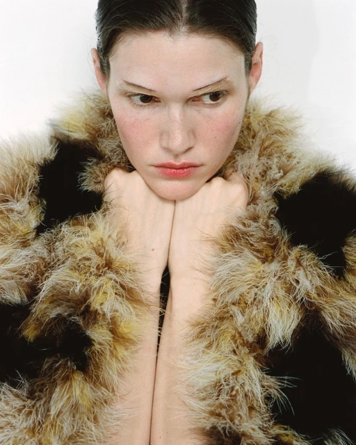 Portrait of a fair-skinned young woman with her chin resting on her hands and wearing a fuzzy, brown and beige faux fur coat in a studio.